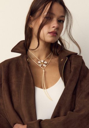 Giovane donna con giacca marrone sopra una maglietta bianca, con una collana a forma di fiore e ciocche di capelli sciolte su uno sfondo uniforme.