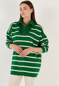 Groen gestreept pullover met een ritskraag. Kenmerken zijn witte horizontale strepen, geribbelde textuur en een ontspannen pasvorm. Lange mouwen.