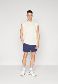 Men’s sleeveless cream shirt with "BJÖRN BORG" logo, paired with navy shorts and white sneakers, set against a plain gray background.