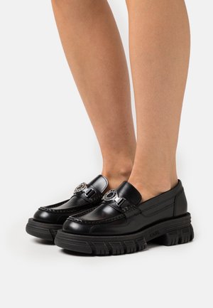 Zwarte leren instappers met glanzende accenten, stevige rubberen zolen, decoratieve metalen gesp en stiksels rondom de rand.