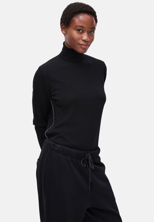 Vrouw met kort krullend haar, gekleed in een zwarte coltrui en zwarte broek met trekkoord, staand tegen een effen lichte achtergrond.