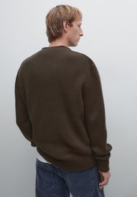Maglione marrone a coste con una vestibilità comoda, dotato di un colletto a girocollo e maniche cucite. Indossato sopra una camicia bianca, abbinato a jeans blu.