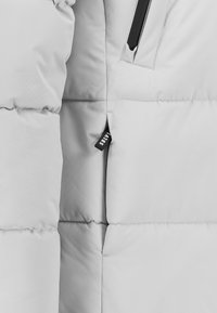 Graue Steppjacke mit einem gesteppte Design, mit einer vertikalen Reißverschlusstasche und einem Etikett an der Seite. Glatte Textur und leichtes Material.