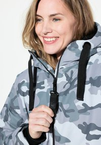 Grå kamouflage vattentät jacka med huva, med dragkedja, svarta dragskor och strukturerad tyg. Närke upp på en person som fäster dragkedjan.