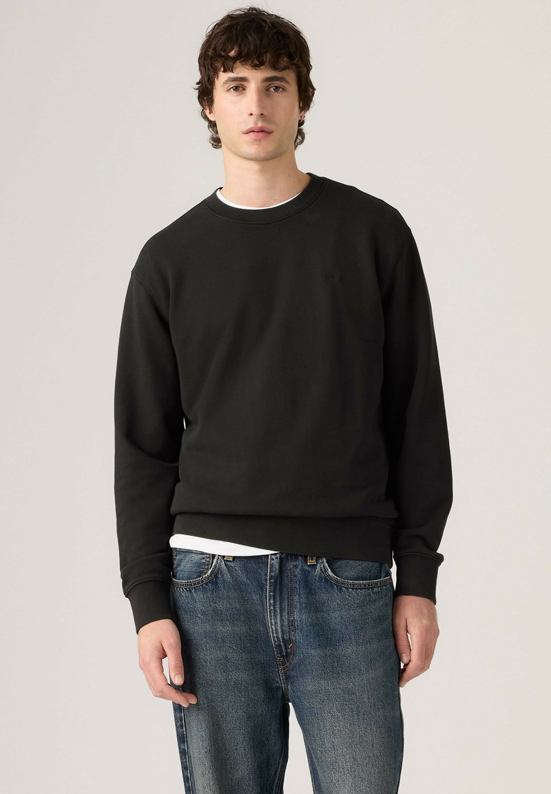 Jeune homme aux cheveux bouclés et foncés porte un sweat-shirt noir sur une chemise blanche et un jean bleu sur un fond clair uni.