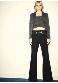 Grauer, kurz geschnittener Pullover mit gerippter Textur, kombiniert mit schwarzen, hoch taillierten Schlaghosen. Silberner Schnallenakzent am Gürtel. Einfacher Hintergrund.