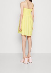 Gul passform-och-utvidgad sommarklänning med smockad överdel, tunna band och en utsvängd kjol, i kombination med rosa blockklackade mule-skor.