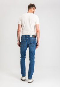 Mężczyzna w białej koszulce, niebieskich dżinsach z metką z tyłu pasa oraz białych trampkach, stojący tyłem na jednolitym tle.