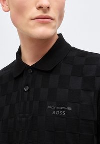 Κοντινό πλάνο ενός ατόμου που φοράει μαύρο καρό πόλο με το λογότυπο "Porsche BOSS" στο στήθος, με ορατά κουμπιά κοντά στον γιακά.