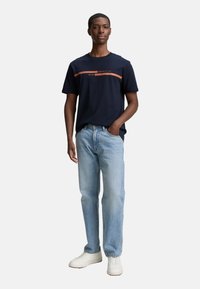 Marineblå bomulds t-shirt med en orange grafisk stribe over brystet, parret med lyseblå jeans med lige ben og hvide sneaks.