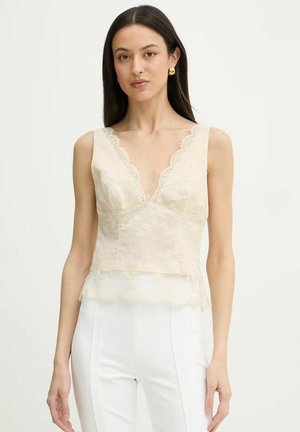 Donna che indossa un top in pizzo beige senza maniche con scollo a V smerlato abbinato a pantaloni bianchi a vita alta, in piedi su sfondo semplice.