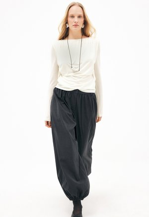 Frau mit langen blonden Haaren trägt ein weißes langärmeliges Oberteil, lockere schwarze Hosen mit elastischen Bündchen, schwarze Schuhe und eine Halskette, steht vor weißem Hintergrund.