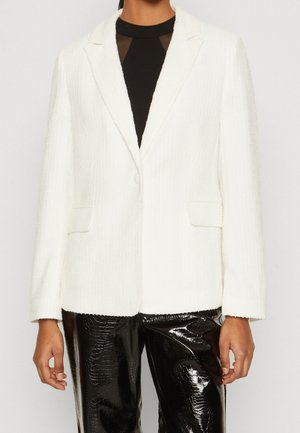 Femme portant un blazer blanc texturé sur un top en maille noire et un pantalon noir en cuir synthétique brillant avec motif crocodile.