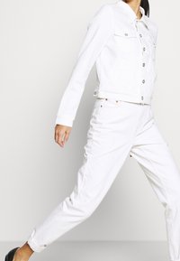 Chaqueta de mezclilla blanca con bolsillos frontales y cierres de botón, combinada con pantalones de mezclilla blanca a juego. Ambos tienen un corte relajado y una textura suave.