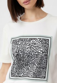 Weißes T-Shirt mit einem großen, schwarz-silbernen gepunkteten Grafik innerhalb eines mehrfarbigen Rahmens. Kurze Ärmel und eine lockere Passform.