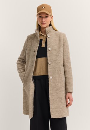Frau trägt einen cremefarbenen, strukturierten Wollmantel mit großen Knöpfen, einen schwarz-creamfarben gestreiften Polo-Pullover, schwarze Hosen und eine beige Baseballmütze.