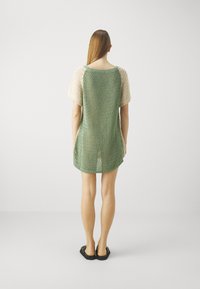Tunique en tricot avec un corps vert et des manches courtes beiges, présentant un design en maille lâche et un ourlet droit. Portée avec des sandales noires.