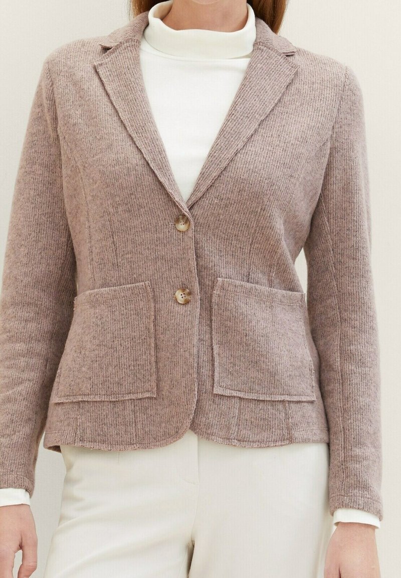 Blazer en maille marron doux avec deux poches avant et deux boutons, porté sur un col roulé blanc et associé à un pantalon crème.