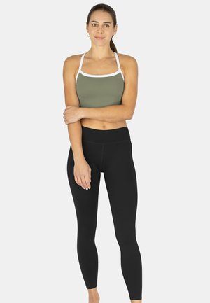 Junge Frau steht mit verschränkten Armen, trägt einen olivgrünen Sport-BH mit weißen Trägern und schwarze, hoch taillierte Leggings vor einem einfarbigen Hintergrund.