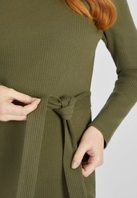 Olivgrünes geripptes Kleid mit langen Ärmeln, das ein gebundenes Taillen-Detail aufweist. Der Stoff wirkt weich mit subtiler Textur und strukturierter Form.