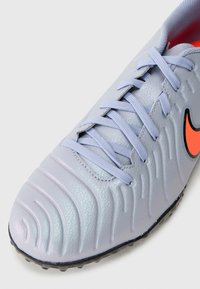 Scarpa da calcio sintetica grigia chiara con superficie testurizzata, lacci viola, suola nera e un accento arancione dello swoosh Nike.