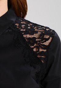 Sort bluse med udsmykket sort spidsel på venstre skulder, glat stof og et klassisk kravedesign.