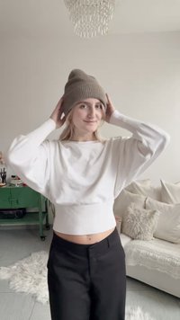 Pull blanc court avec ourlet côtelé, manches longues bouffantes, et un bonnet tricoté marron. Pantalon noir ; en arrière-plan, des textiles clairs et un mobilier vert.