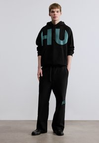Μαύρο φούτερ με κουκούλα με μεγάλο τυρκουάζ λογότυπο "HU", τσέπη καγκουρό μπροστά, συνδυασμένο με μαύρο παντελόνι φόρμας με μικρότερο τυρκουάζ "HU" στο πόδι.