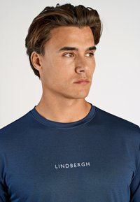 Ung mand med kort brunt hår iført en marineblå Lindbergh skjorte, der kigger til højre mod en ensfarvet hvid baggrund.