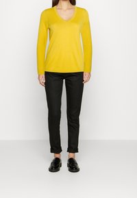 Geel V-hals trui met lange mouwen, gecombineerd met zwarte slim-fit broek opgerold bij de boorden, en zwarte instappers, gepresenteerd tegen een neutrale achtergrond.