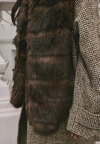Brązowa kamizelka z futra ekologicznego nałożona na płaszcz w kratę houndstooth w odcieniach beżu i czerni, prezentująca teksturowane, miękkie wykończenie i luźny krój.
