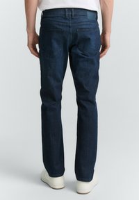 Hombre vestido con jeans rectos de color azul oscuro y zapatillas blancas, de pie con las manos relajadas a los lados frente a un fondo simple.