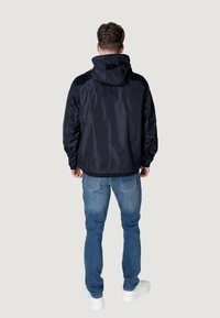 Giacca impermeabile blu navy con cappuccio, polsini elastici e vestibilità comoda, abbinata a jeans blu e sneakers bianche. Texture liscia, senza fantasie.