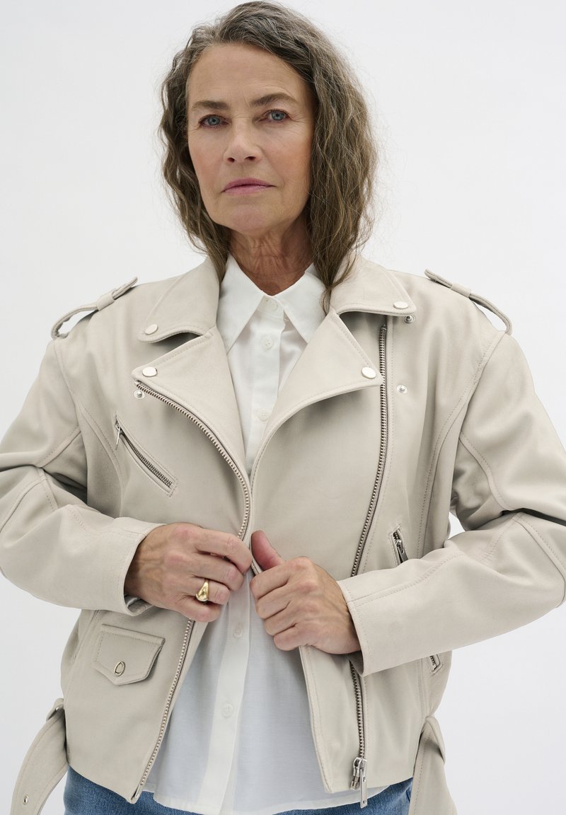 Beige Kunstleder-Motorradjacke mit asymmetrischem Reißverschluss, Reißverschlusstaschen und Schulterepauletten. Getragen über einer weißen Bluse.