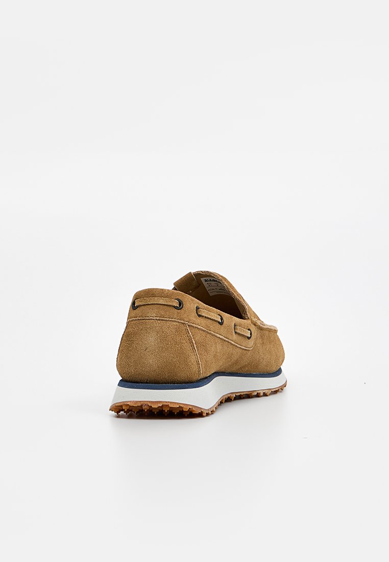 Bateau en suède beige avec une forme arrondie, doté d'une semelle blanche et bleue, d'œillets décoratifs et d'une semelle extérieure texturée pour une adhérence optimale.