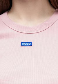 Jasnoróżowy prążkowany t-shirt z niebieską, prostokątną naszywką z logo HUGO na piersi. Dekolt okrągły, z widocznymi przeszyciami.