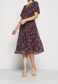 Černé květinové šaty s balónovými rukávy, výstřihem do V a splývavou midi délkou. Vzor obsahuje růžové a žluté květiny na průhledné látce. Béžové sandály na vysokém podpatku.