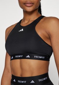 Svart sports-BH laget av elastisk materiale, med høy hals, racerback-design og hvit "TECHFIT"-merkevare langs båndet.