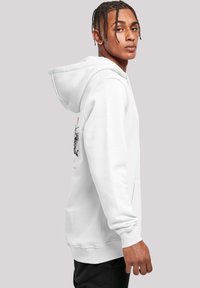 Young man with braided hair wearing a white hoodie with a black and orange graphic on the back, standing against a plain background.