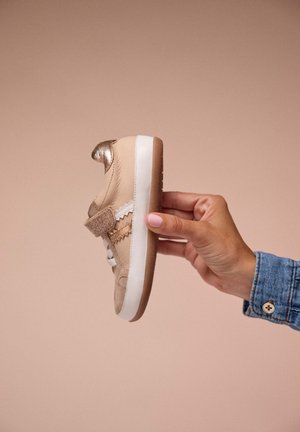 Baskets beiges en daim texturé avec des accents dorés brillants. Présentent une bande Velcro et une semelle en caoutchouc blanc. Tenues contre un fond rose doux.