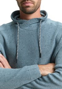 Blauer strukturierter Pullover mit hohem Kragen, ausgestattet mit einem Kordelzug mit schwarzen Spitzen und einem gerippten Muster. Der Stoff enthält Farbspritzer.