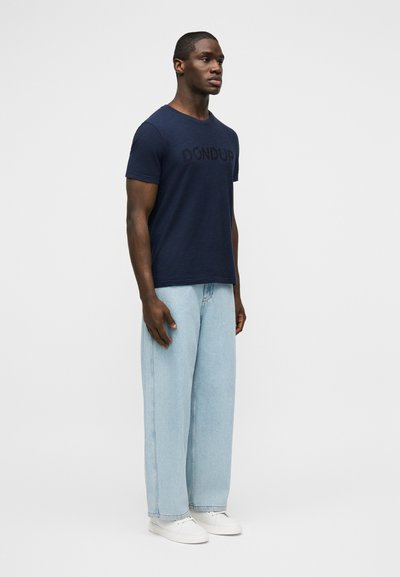 Άνδρας που στέκεται προς τα εμπρός φορώντας μπλε σκούρο μπλουζάκι με το κείμενο "DONDUP", ανοιχτόμπλε παντελόνι με φαρδιά μπατζάκια και λευκά αθλητικά παπούτσια.