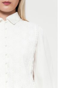 Hvit bluse med krage, med blomsterbroderi på forsiden og bølget kant. Knapper løper nedover midten. Lange ermer.