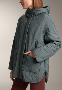 Quilted, hooded jacket in dark green with oversized fit. Features side pockets and a high collar, made from a smooth, lightweight material.