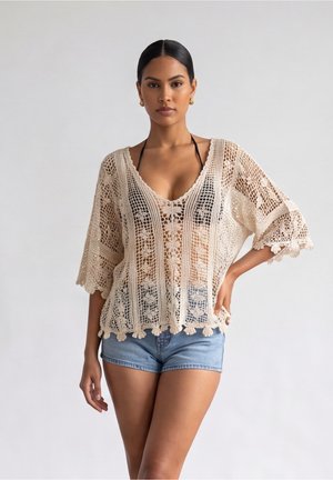 Donna che indossa un top beige all'uncinetto sopra un top bikini nero e pantaloncini di jeans, in piedi con una mano sui fianchi davanti a uno sfondo semplice.