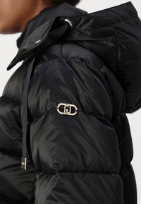 Jachetă puf de culoare neagră, cu un material neted și lucios, glugă cu șnururi și un accent metallic al logo-ului pe umăr.