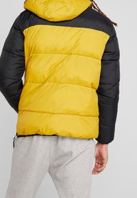 Fodrad jacka med en split-design i stark gul och svart, med huva, dragkedjor på sidorna och horisontell sömnad.