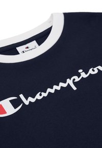 Camiseta de cuello redondo en color marino con un cuello de canalé blanco y un gran logotipo blanco de "Champion"; la tela es de algodón suave con una textura lisa.