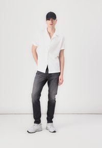 Chemise blanche à manches courtes, jeans foncés et baskets blanches. Casquette noire sur la tête. Arrière-plan neutre. Design simple, style décontracté.