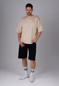 Beige oversized t-shirt with short sleeves, black shorts, and white sneakers, complemented by white socks with "ALCA" printed in black.